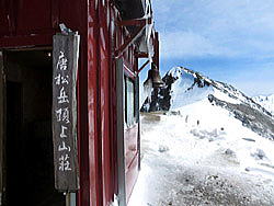 唐松岳頂上山荘の玄関と牛首。稜線は穏やかな晴天の朝です。本日は気温が低く、屋根や鐘に付いたつららは、午後まで解けずに残っていました。